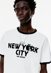 Hvid t-shirt med sort rund hals og ærmer, der har fed tekst "NEW YORK CITY" og et grafisk motiv af Frihedsgudinden. Sølv halskæde som udsmykning.