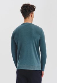 Mężczyzna z krótkimi kręconymi włosami, ubrany w turkusowy sweter z długim rękawem i ciemne spodnie, stoi tyłem na jednolitym tle.