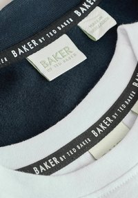 Divas divi krekli, viens tumši zils un otrs balts. Abiem ir auduma birkas ar "BAKER by TED BAKER" kontrastējošās krāsās un tekstūrās.