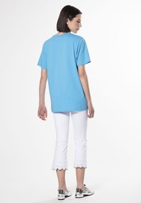 Camiseta de algodón azul de manga corta combinada con pantalones blancos recortados con detalles de ojales en el dobladillo. El modelo está de espaldas, luciendo zapatos negros y plateados.