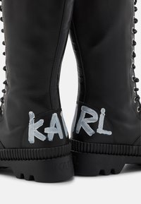 Černé kožené šněrovací boty s tlustými podrážkami, bílý nápis "KARL" natřený na zadní straně paty každé boty.