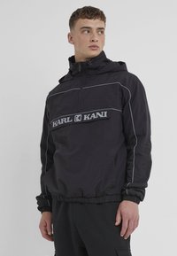 Black hooded pullover jacket made of lightweight fabric, featuring a front zipper, elastic cuffs, and a prominent gray logo band across the chest.