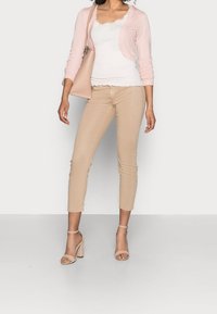 Ljust rosa cardigan över en vit tanktop med spetsdetaljer, kombinerad med beige åtsittande byxor och nakna högklackade sandaler. Enkelt mönster och färger.