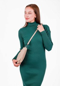 Zelené žebrované šaty s rolákem. V jedné ruce je držena béžová texturovaná kabelka s dlouhým popruhem. Bílé pozadí.