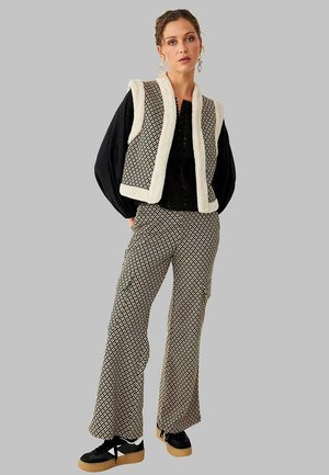 Mujer que lleva pantalones negros con estampado y chaleco sin mangas a juego con ribete blanco, blusa negra de manga larga y zapatillas negras con plataforma.