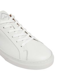 Zapatilla de piel blanca con punta redonda, cordones blancos planos y suela de goma texturizada. Presenta un diseño minimalista y un branding sutil.