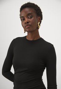 Melnas garās piedurknes blūze ar gludu, elastīgu tekstūru. Modelis valkā zelta abstraktas auskarus. Vienkāršs apaļš kakla izgriezums un pieguļošs dizains.