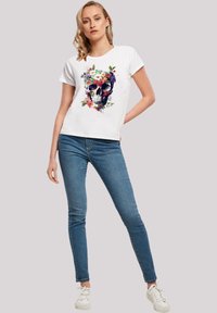 White t-shirt featuring a vibrant floral skull graphic, short sleeves, relaxed fit, paired with blue skinny jeans and white sneakers.