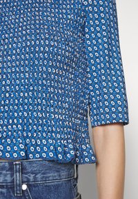 Blouse smockée à motifs bleus avec manches 3/4 portée avec un jean en denim bleu, montrant la texture et le design détaillés du tissu.