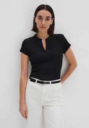 Vrouw met donker schouderlang haar die een zwart topje met korte mouwen en een witte broek met een zwarte riem draagt, staand tegen een witte achtergrond.