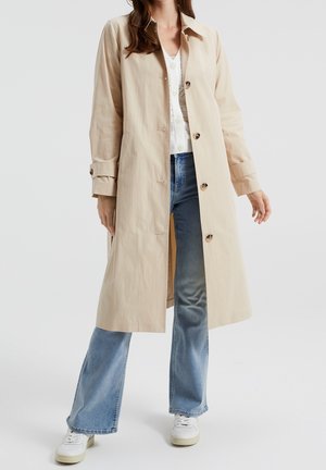 Femme portant un manteau long beige, une chemise blanche, un jean bleu et des baskets blanches, debout devant un fond uni.