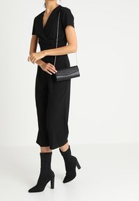 Combinaison noire avec un corsage ajusté, des manches courtes et des jambes larges. Assortie à des bottines noires et un petit sac bandoulière noir avec une chaîne.