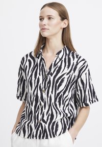 Camicia a maniche corte con una fantasia a strisce zebrate bianche e nere. Presenta un'apertura frontale con bottoni e una vestibilità rilassata con colletto.