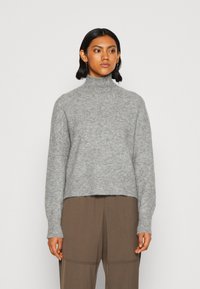 Pull léger gris clair à manches longues en tissu doux et texturé, avec un col haut et un ourlet court, associé à un pantalon brun ample.