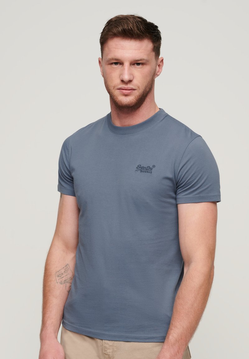 Camiseta de algodón azul, de manga corta, con cuello redondo, que presenta un pequeño logo bordado en la zona izquierda del pecho. Textura suave, diseño ajustado.