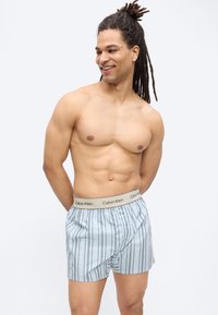 Γυμνός άνδρας με κοτσίδες, φορώντας ανοιχτό μπλε ριγέ μποξεράκια Calvin Klein, χαμογελαστός και κοιτάζοντας προς τα αριστερά, σε απλό φόντο.