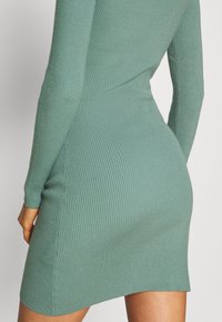 Robe côtelée bleu sarcelle avec des manches longues, présentant une silhouette ajustée et une fente sur le côté, fabriquée en maille douce.