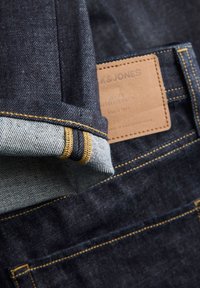 Tumesinised teksapüksid, millel on kuldsed õmblused ja nahast plaastriga logo. Servad on ülespoole keritud, paljastades heledama riide all.