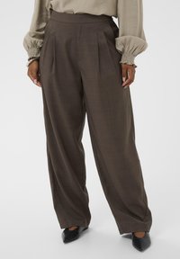 Laia püksid tumepruunist kangast, vertikaalsete voldikute ja peene tekstuuriga. Sobitatud mustade terava ninaga kingadega.