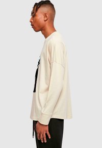 Beige Langarm-T-Shirt mit Rundhalsausschnitt, mit einem grafischen Druck auf der Vorderseite. Aus Baumwolle hergestellt, hat einen lockeren Schnitt und eine dezente Textur.