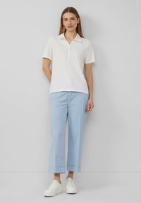 Weißes Kurzarm-Polohemd mit Kragen, kombiniert mit hellblauen, schmal geschnittenen, cropped Hosen und weißen Sneakern vor einem einfarbigen Hintergrund.