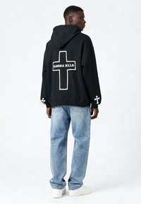 Felpa nera con testo bianco "KARMA KILLS" e design a croce sul retro; polsini a coste; abbinata a jeans blu chiaro a vestibilità rilassata.