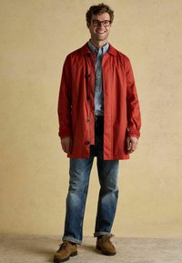 Raudona vandeniui atspari striukė su juodomis sagomis, dėvima ant mėlynos ir baltos ruoželiais marškinėlių; derinama su nudėvėtais mėlynais džinsais ir rudais batais.