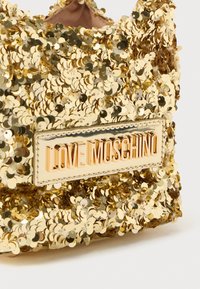 Zlata torbica pokrita s kosmiči s pravokotno kovinsko ploščico, na kateri so izbočene črke "LOVE MOSCHINO".