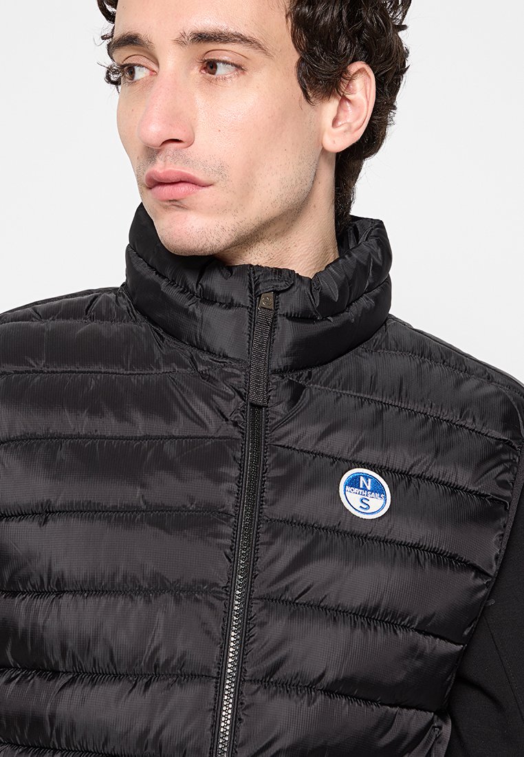 Gilet imbottito nero con colletto alto, texture trapuntata e chiusura a zip. Presenta una patch con logo blu e bianco sul lato sinistro.