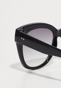 Óculos de sol pretos com acabamento brilhante, forma de gato, lentes em degradê e pontos de realce dourados na haste. Design sutil e moderno.