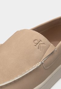 Μπεζ παπούτσι τύπου slip-on με λευκή σόλα, με ανάγλυφο λογότυπο "ck" στο πάνω μέρος και ορατές λευκές ραφές κατά μήκος της μπροστινής άκρης.