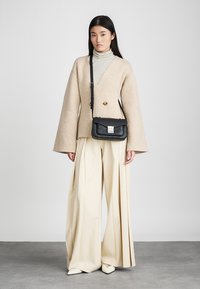 Beigā vilnas maisījuma apvalks ar platām piedurknēm, kombinēts ar strīpota augstās kakla krekla, krēma svītrainām biksēm un melnu crossbody somu.