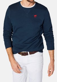 Granatowy bawełniany sweatshirt z długimi rękawami i okrągłym dekoltem. Posiada czerwone haftowane logo na piersi. Noszony z białymi spodniami i brązowym paskiem.
