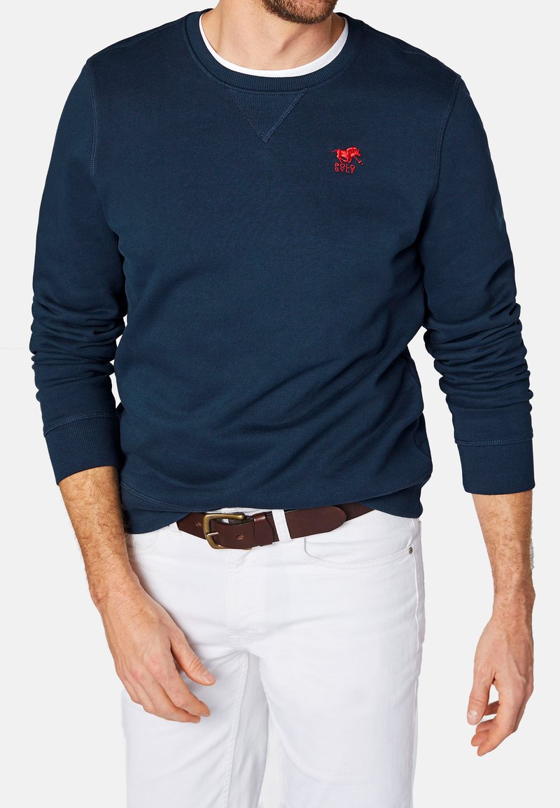 Granatowy bawełniany sweatshirt z długimi rękawami i okrągłym dekoltem. Posiada czerwone haftowane logo na piersi. Noszony z białymi spodniami i brązowym paskiem.