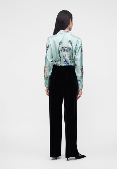 Femme debout, vue de dos, portant une chemise en satin bleu clair avec des motifs imprimés et un pantalon en velours noir taille haute avec des talons.