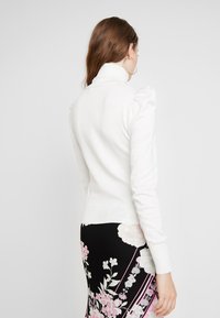 Weißer, figurbetonter, langärmeliger Rollkragenpullover mit Puffdetails an den Schultern. Glatte Textur; kombiniert mit einem floral gemusterten Rock in Schwarz und Pink.