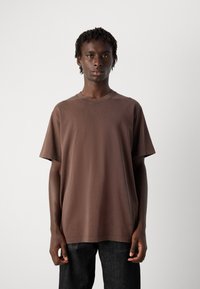 Young man with short dreadlocks wearing a plain brown oversized t-shirt and dark pants, standing against a plain light background.