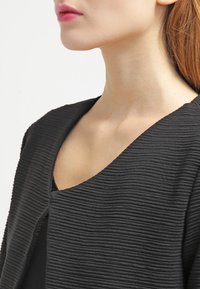 Svart texturerad jacka med ett ribbad mönster och bred V-nick, med en figurnära silhuett och slät tyg.