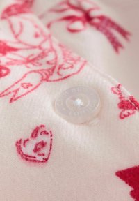 Przezroczysty guzik przyszyty do miękkiej białej tkaniny z czerwonym sercem i ilustracjami zwierząt, na guziku znajduje się napis "CATIM KIDS LONDON".