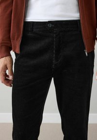 Black corduroy pants feature a ribbed texture, front button closure, and pockets, paired with a red zippered jacket.