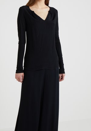 Kobieta ubrana w czarną bluzkę z długim rękawem i dekoltem w serek oraz szerokie czarne spodnie, stojąca na tle jednolitego jasnoszarego tła.