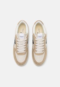 Beżowe i kremowe sneakersy z materiałów zamszowych i skórzanych, perforowanym noskiem, białymi sznurówkami oraz czarnymi akcentami logo.
