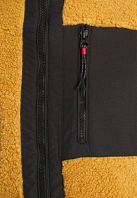 Giacca in pile con tessuto giallo testurizzato sopra, materiale nero liscio sotto e due tasche con zip con accenti rossi.