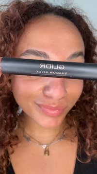 Femme aux cheveux bouclés tient un GLIDR Shadow Stick noir horizontalement devant ses yeux, souriant doucement.