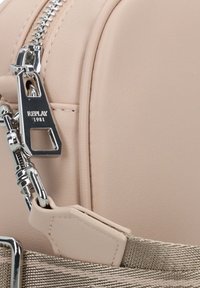 Dettaglio di uno zaino sintetico beige con cerniera e hardware argentati, con una tracolla testurizzata e un'accents del logo. Superficie liscia con cuciture.