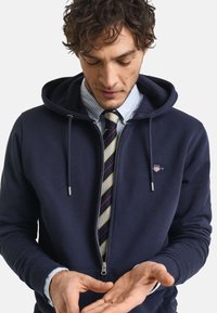 Navy zip-up hoodie made of soft fabric, featuring drawstrings, ribbed cuffs, and a small logo. Worn over a light blue striped shirt with a tie.