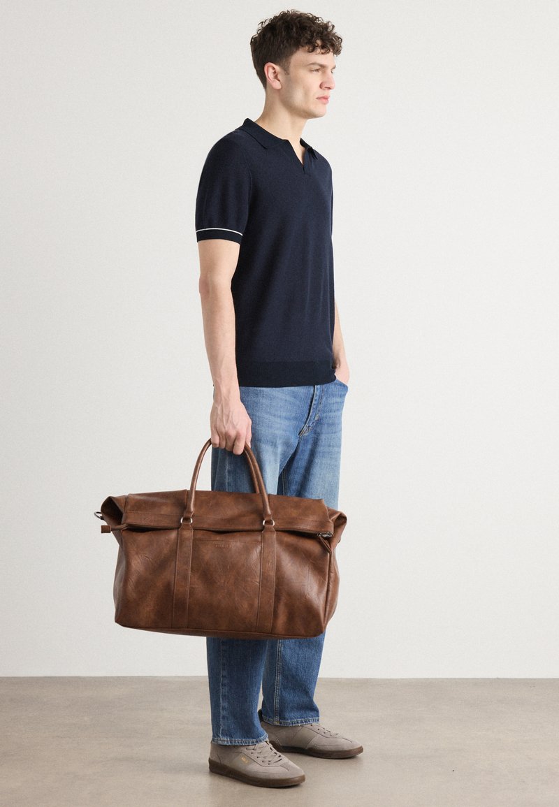 Sac de voyage en cuir marron avec une texture lisse, taille grande et deux poignées courtes, présenté sur un fond uni avec une personne le tenant.