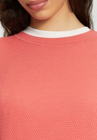 Korallenfarbener Pullover mit Rundhalsausschnitt und kontrastierendem weißen Innenkragen. Der Stoff weist ein Wabenmuster auf.