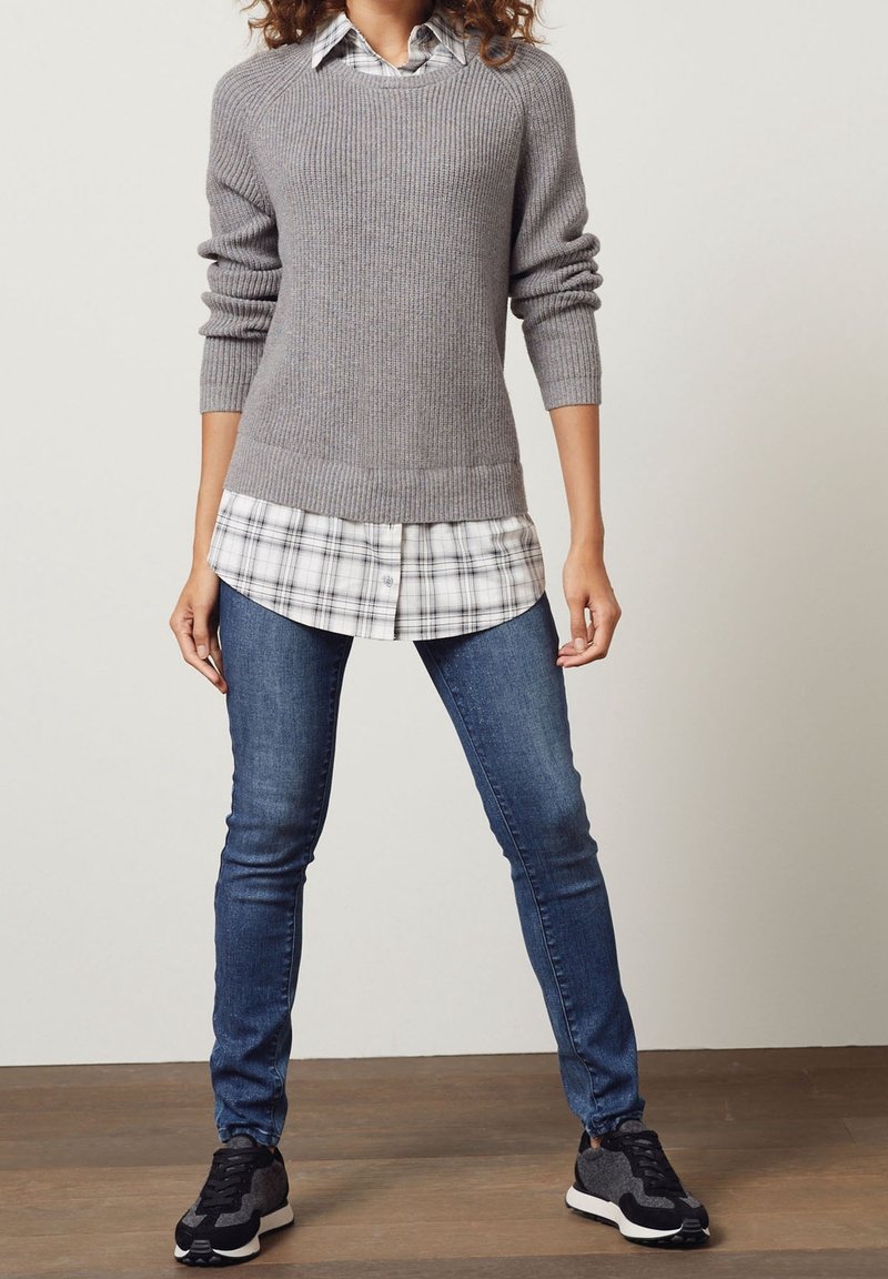 Femme portant un pull en maille gris par-dessus une chemise blanche à carreaux, un jean skinny bleu et des baskets noires, debout devant un fond uni.