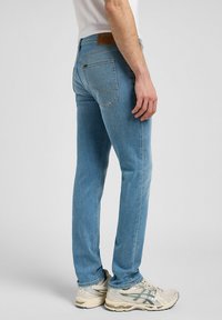 Mann iført lyseblå rette jeans og hvite joggesko, stående sideveis mot en ensfarget grå bakgrunn.